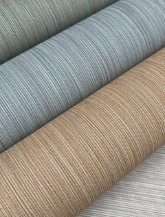 Three rolls of York Wallcoverings’ Cannete Wallpaper, featuring blue, beige, and brown tones with subtle horizontal lines, are stacked closely together on a light-colored surface.
