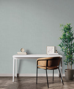 A white desk with a clock, books, and framed art sits against a wall covered in light blue, textured Magnolia Home Meadowbrook Wallpaper.