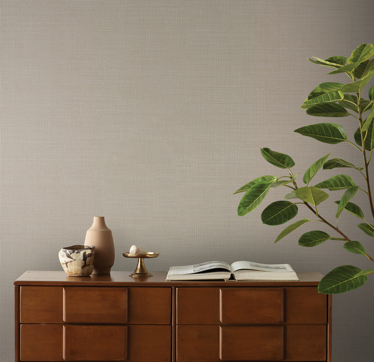 Light gray Magnolia Home Meadowbrook Wallpaper by York Wallcoverings with a fine, textured weave pattern behind a wooden dresser holding decorative objects and an open book.