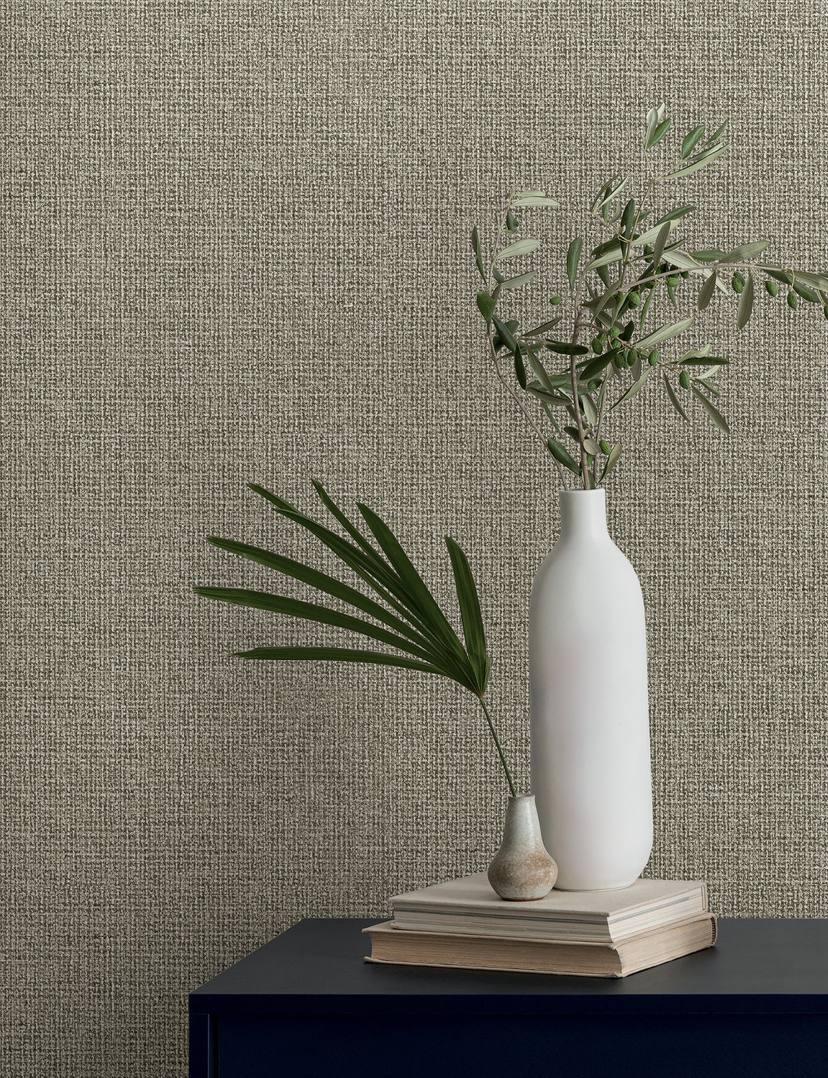 Two vases with leafy branches rest on a stack of books against a textured, neutral-toned wall covered in Magnolia Home Brindled Wallpaper by York Wallcoverings.