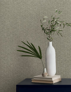 Two vases with leafy branches rest on a stack of books against a textured, neutral-toned wall covered in Magnolia Home Brindled Wallpaper by York Wallcoverings.