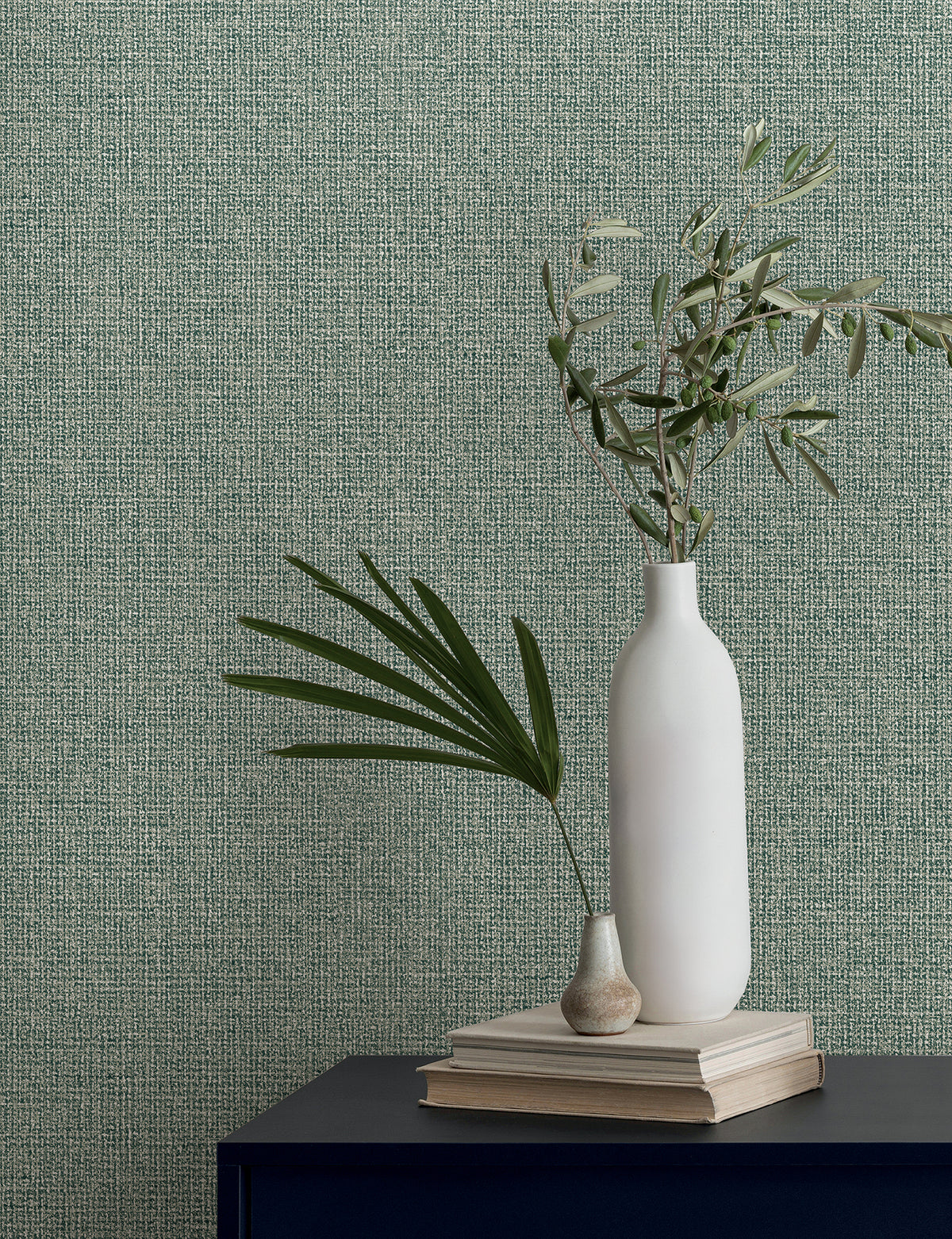 Two vases holding leafy branches sit on a stack of books against green textured Magnolia Home Brindled Wallpaper.