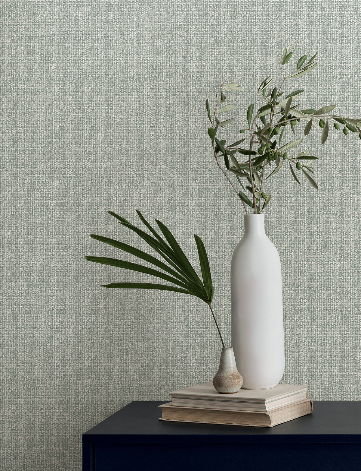 Magnolia Home Brindled Wallpaper in light green with a textured, woven pattern, shown behind a shelf with two vases and olive branches on a stack of books.