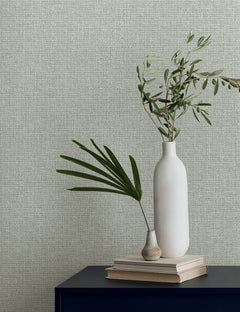 Magnolia Home Brindled Wallpaper in light green with a textured, woven pattern, shown behind a shelf with two vases and olive branches on a stack of books.