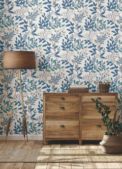 A wooden dresser and floor lamp stand on a wooden floor against a white wall featuring blue and gray leafy floral Eden Retreat Wallpaper by York Wallcoverings. A potted plant and wicker baskets sit atop the dresser.