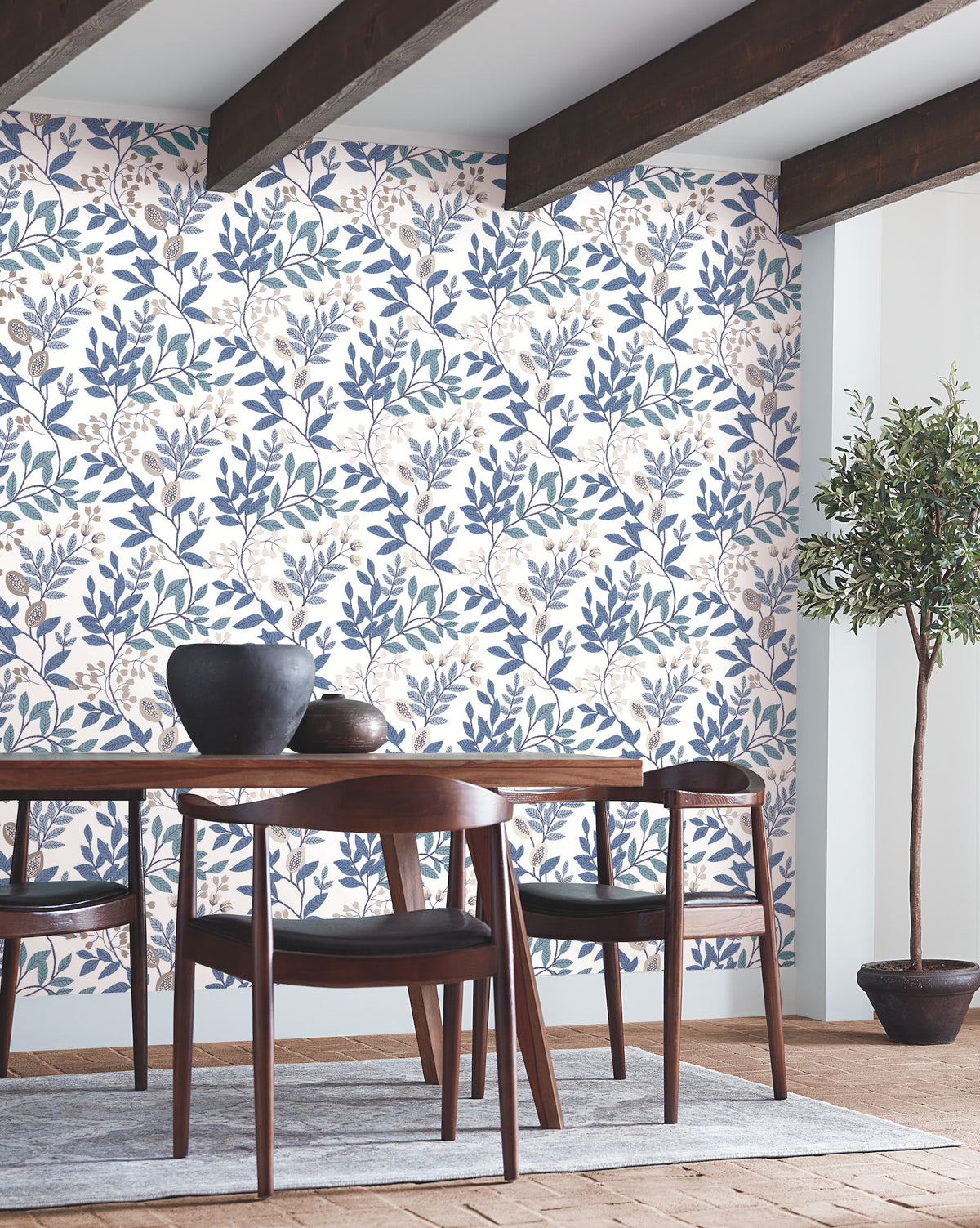 Dining room featuring a wooden table and chairs, decorative bowls on the table, Eden Retreat Wallpaper by York Wallcoverings in a blue and beige floral pattern, exposed ceiling beams, hardwood floor, and a potted tree in the corner.