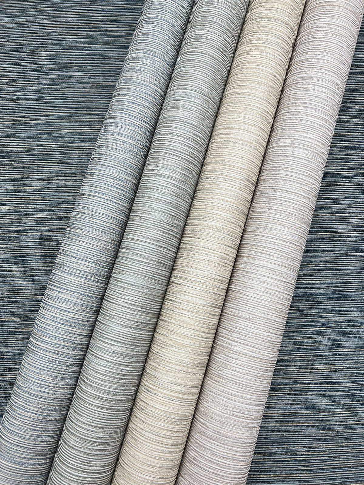 Four rolls of Grass Roots Wallpaper featuring textured, horizontally-striped patterns in shades of dark gray, light gray, beige, and cream.