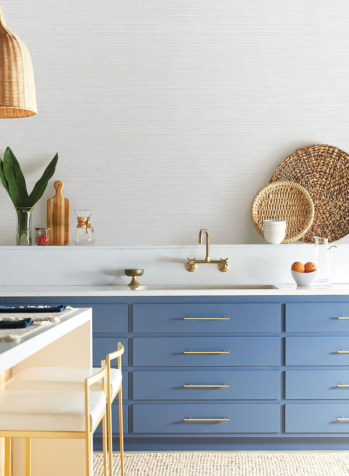 Blue kitchen cabinets with gold hardware, a wall-mounted brass faucet, and neutral textured Grass Roots Wallpaper by York Wallcoverings.
