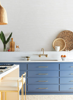 Blue kitchen cabinets with gold hardware, a wall-mounted brass faucet, and neutral textured Grass Roots Wallpaper by York Wallcoverings.