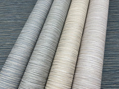 Four rolls of Grass Roots Wallpaper in shades of gray, beige, and taupe displayed diagonally against a dark gray horizontal-lined background.