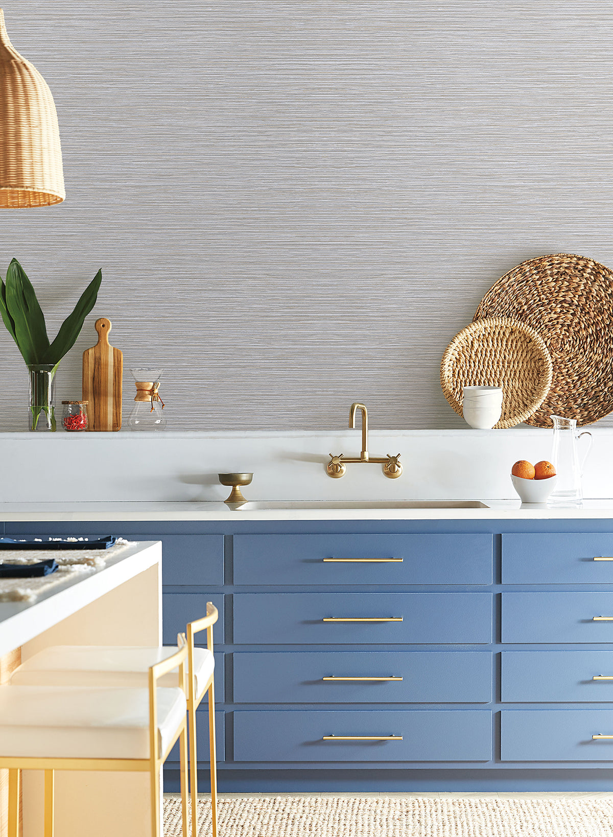 A kitchen sink area features blue cabinets and walls covered in light-colored, horizontal-textured Grass Roots Wallpaper by York Wallcoverings.