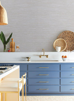A kitchen sink area features blue cabinets and walls covered in light-colored, horizontal-textured Grass Roots Wallpaper by York Wallcoverings.