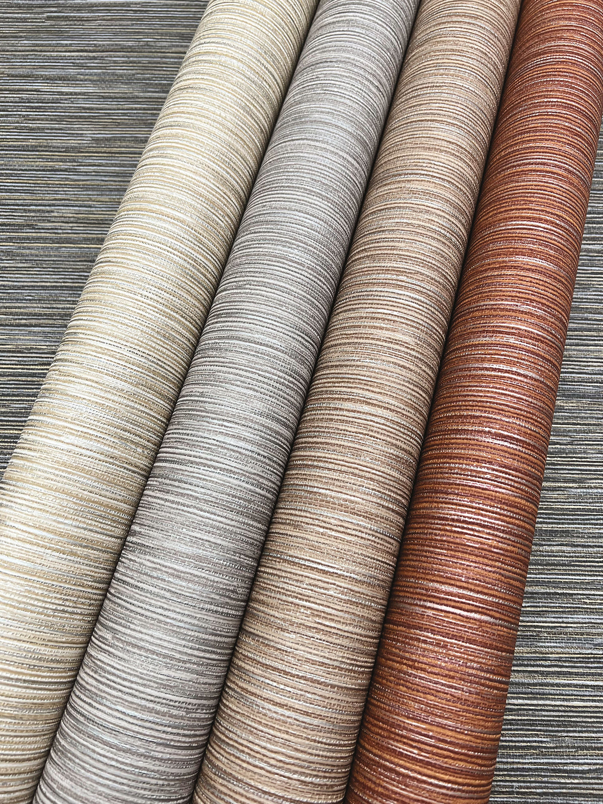 Four rolls of Grass Roots Wallpaper by York Wallcoverings in varying shades of cream, gray, beige, and copper laid diagonally against a dark gray background.