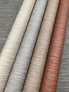 Four rolls of Grass Roots Wallpaper by York Wallcoverings in varying shades of cream, gray, beige, and copper laid diagonally against a dark gray background.