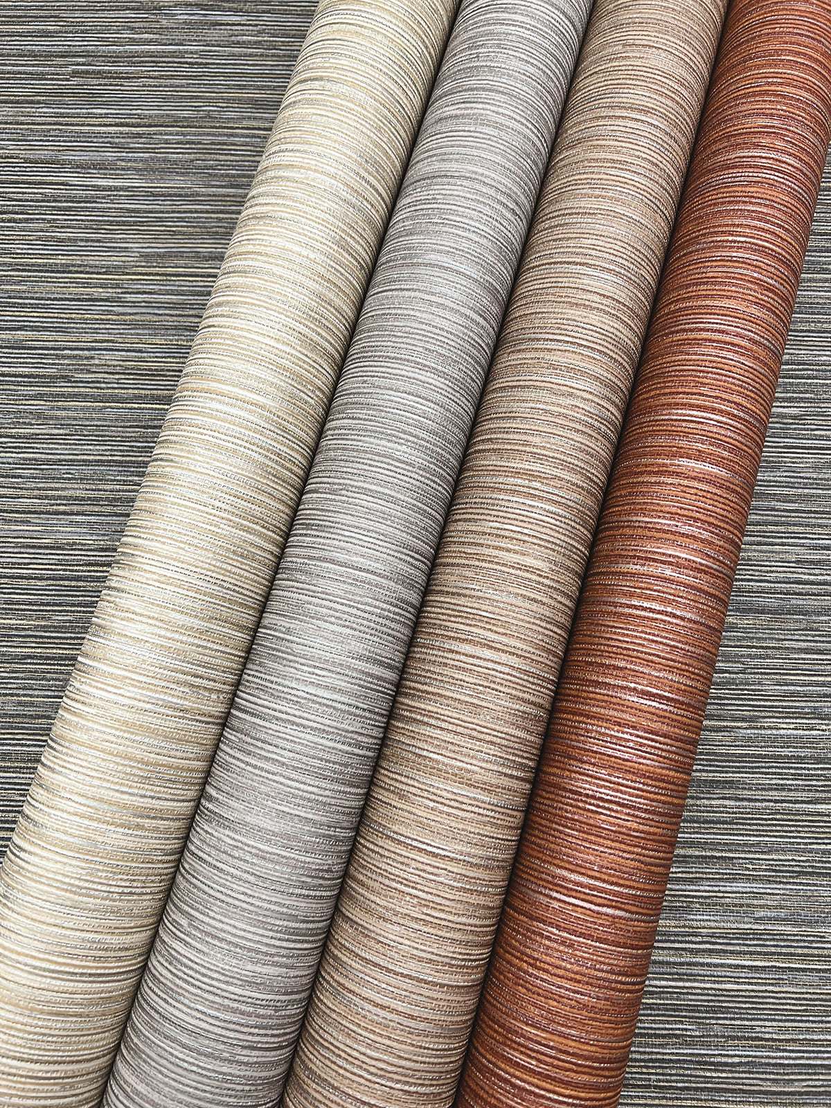 Four rolls of Grass Roots Wallpaper in cream, silver, taupe, and copper displayed diagonally over a dark textured background.