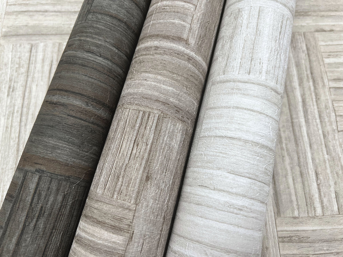 Three rolls of Tesselle Wallpaper featuring a wood-grain geometric pattern in charcoal, taupe, and light gray.