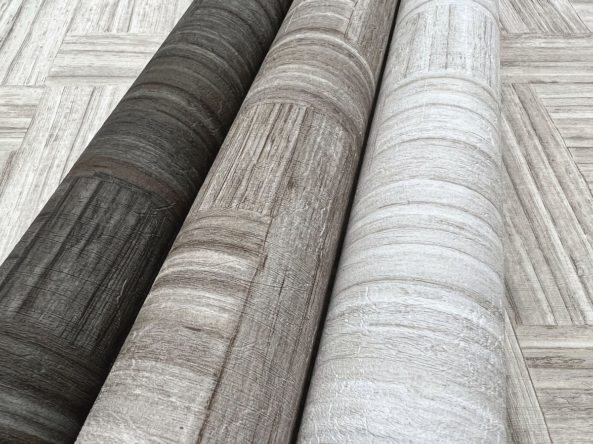 Three rolls of Tesselle Wallpaper in charcoal, taupe, and light gray, each featuring a textured wood-plank pattern.