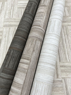 Three rolls of Tesselle Wallpaper featuring a wood-grain geometric pattern in shades of charcoal, taupe, and off-white.