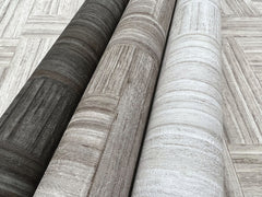 Three rolls of Tesselle Wallpaper featuring a wood-grain pattern in charcoal, brown, and light gray, displayed against a background of the same pattern.