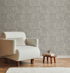 A neutral armchair sits next to a small wooden stool with books and a mug, set against York Wallcoverings Tesselle Wallpaper featuring a repeating geometric wood-grain pattern.