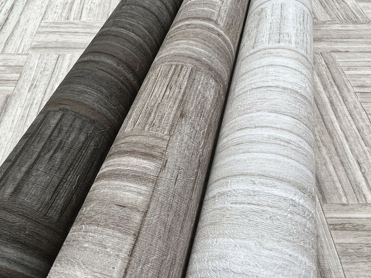 Three rolls of Tesselle Wallpaper in dark charcoal, light brown, and off-white featuring a textured wood-grain pattern, lying on a surface with the same geometric wood-plank design.