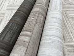 Three rolls of Tesselle Wallpaper in dark charcoal, light brown, and off-white featuring a textured wood-grain pattern, lying on a surface with the same geometric wood-plank design.