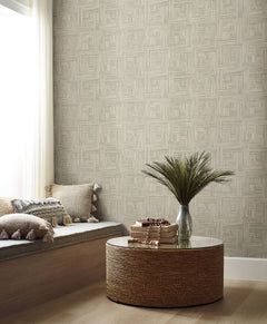 Tesselle Wallpaper by York Wallcoverings features a geometric pattern of overlapping light brown rectangular blocks on a wall behind a cozy seating area and a round woven coffee table.