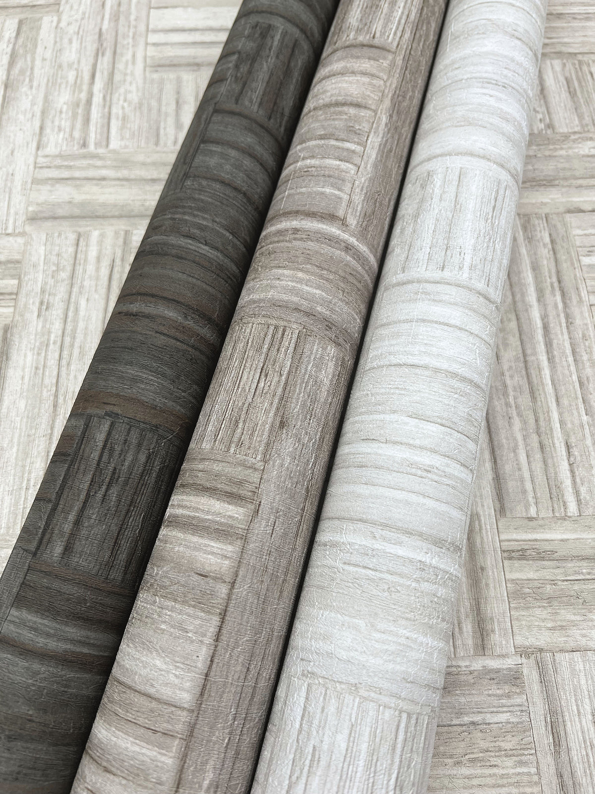 Three rolls of Tesselle Wallpaper in dark gray, beige, and white, each featuring a textured, woven geometric pattern.