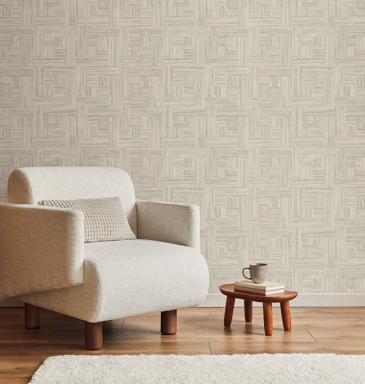 Tesselle Wallpaper by York Wallcoverings in a beige, geometric, woven-look pattern installed behind an armchair, a small side table, and a wooden floor.