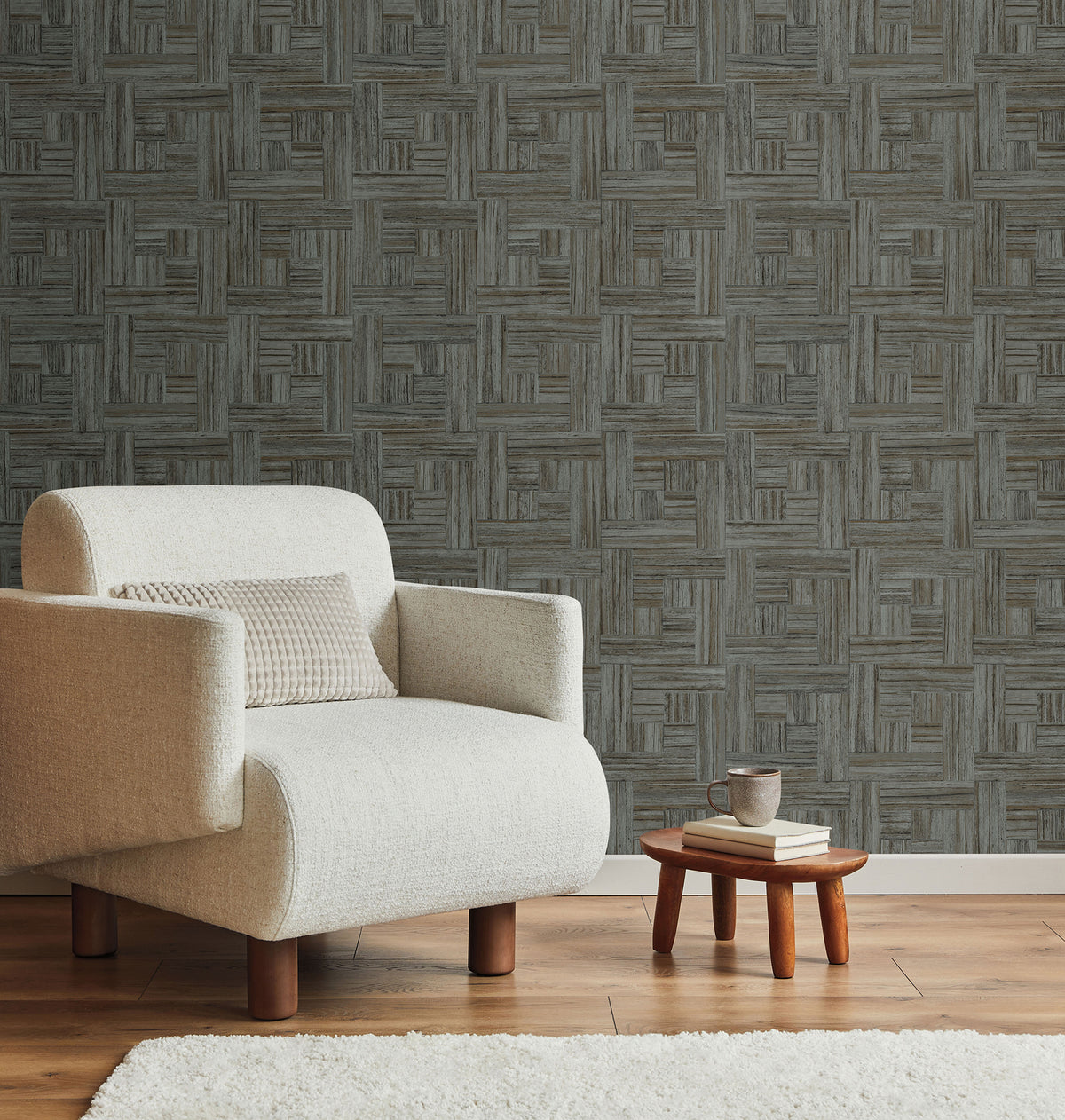 Tesselle Wallpaper featuring a geometric pattern of dark gray and brown wood-grain tiles, shown behind a white armchair and a small side table.