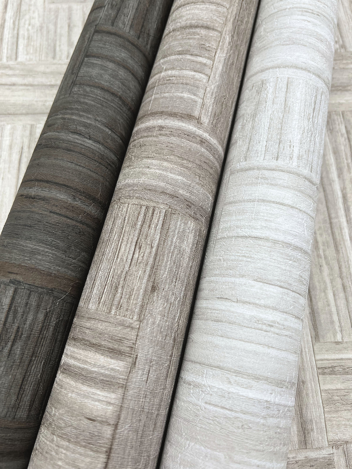 Three rolls of Tesselle Wallpaper in dark gray, brown, and light gray, each featuring a textured, geometric wood-grain pattern.