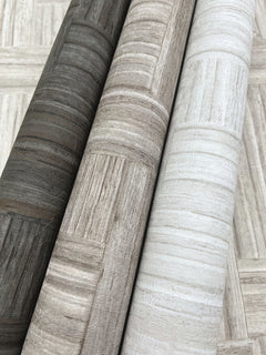 Three rolls of Tesselle Wallpaper in dark gray, brown, and light gray, each featuring a textured, geometric wood-grain pattern.