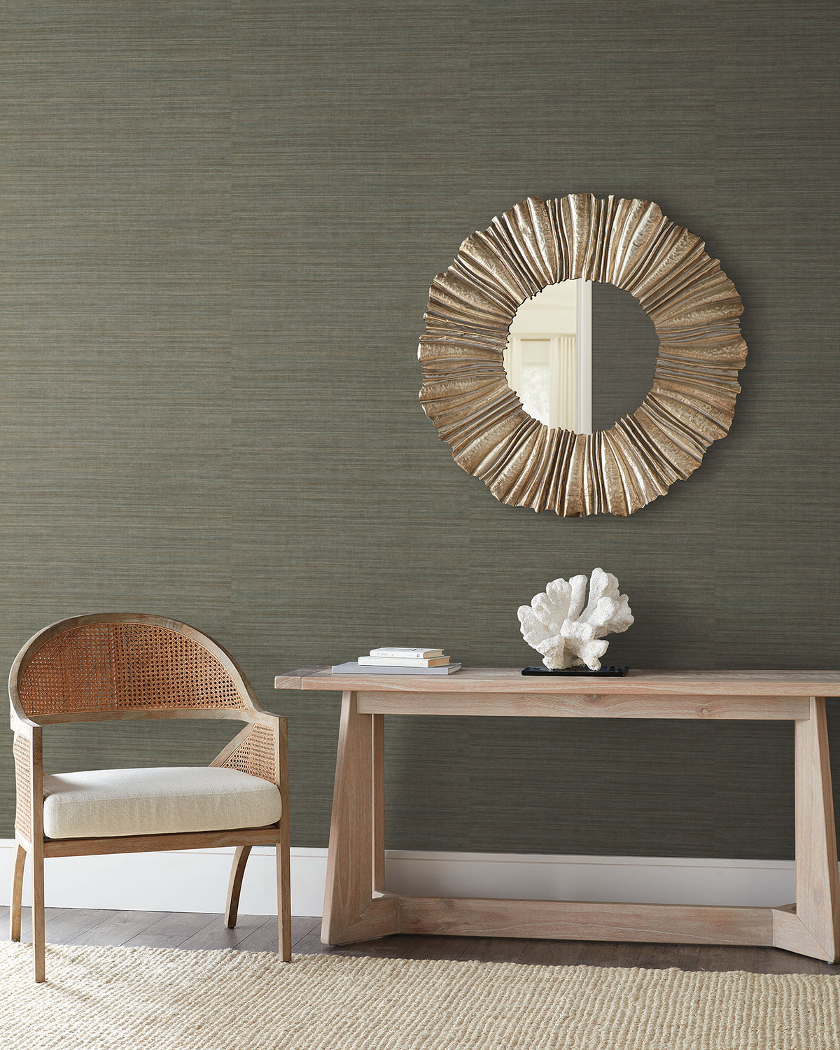 Dark, textured Tasar Silk Wallpaper by York Wallcoverings behind a wooden console table with a coral sculpture, a circular sunburst mirror, and a cane-backed chair.