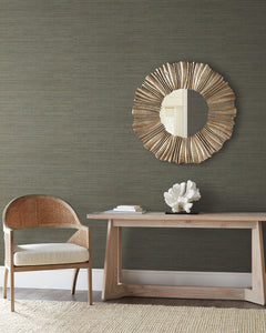 Dark, textured Tasar Silk Wallpaper by York Wallcoverings behind a wooden console table with a coral sculpture, a circular sunburst mirror, and a cane-backed chair.
