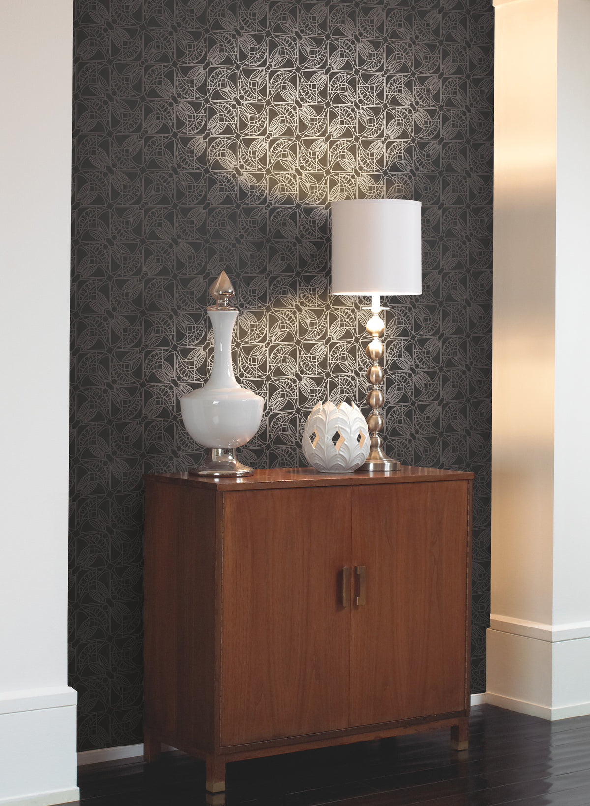 Dark grey Cartouche Wallpaper with a repeating geometric fan pattern, featuring a wooden cabinet topped with a table lamp and two decorative vessels.