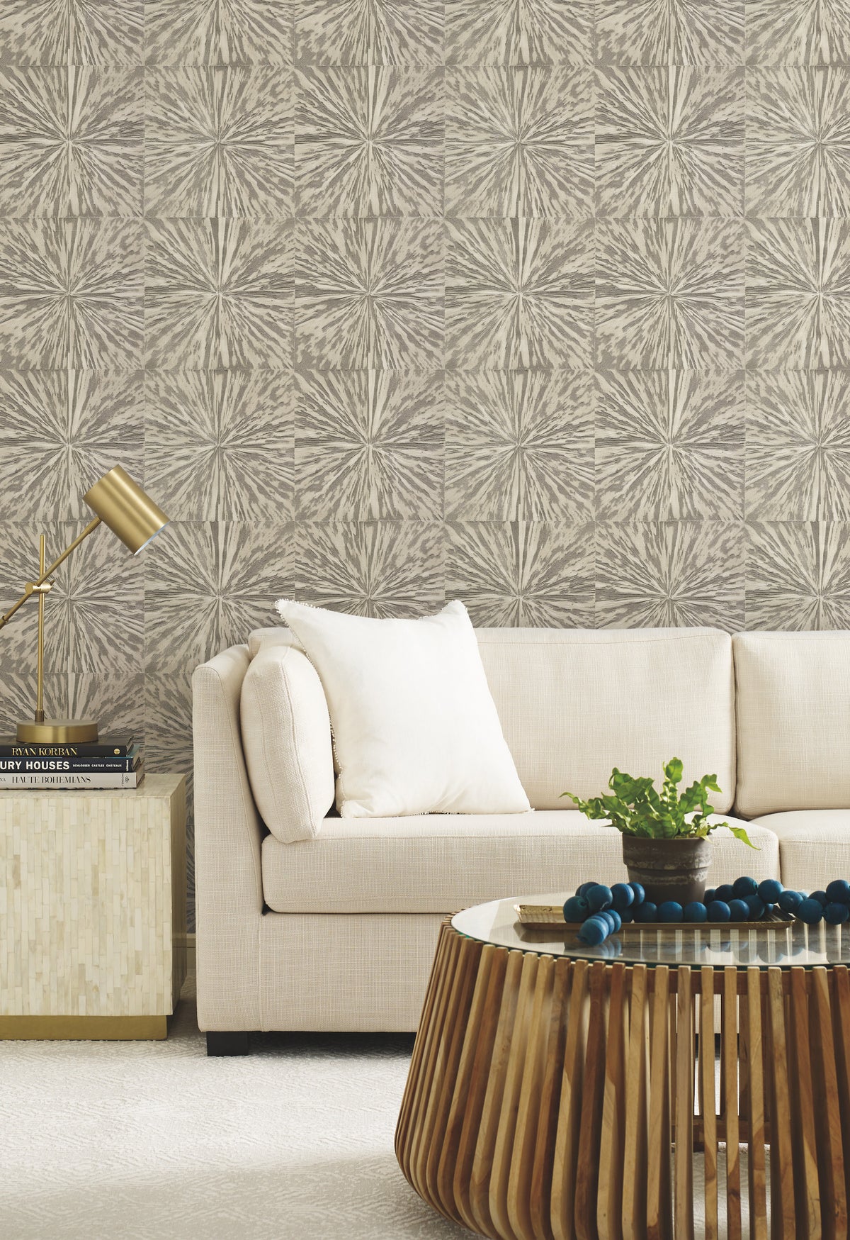 Modern living room with a beige sofa, white pillow, gold lamp on a side table, and a wooden coffee table adorned with beads and a green plant. The wall features Squareburst Wallpaper in neutral tones by York Wallcoverings.