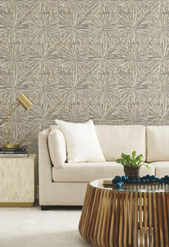 Modern living room with a beige sofa, white pillow, gold lamp on a side table, round wooden coffee table with decor, and York Wallcoverings’ Squareburst Wallpaper in neutral tones.