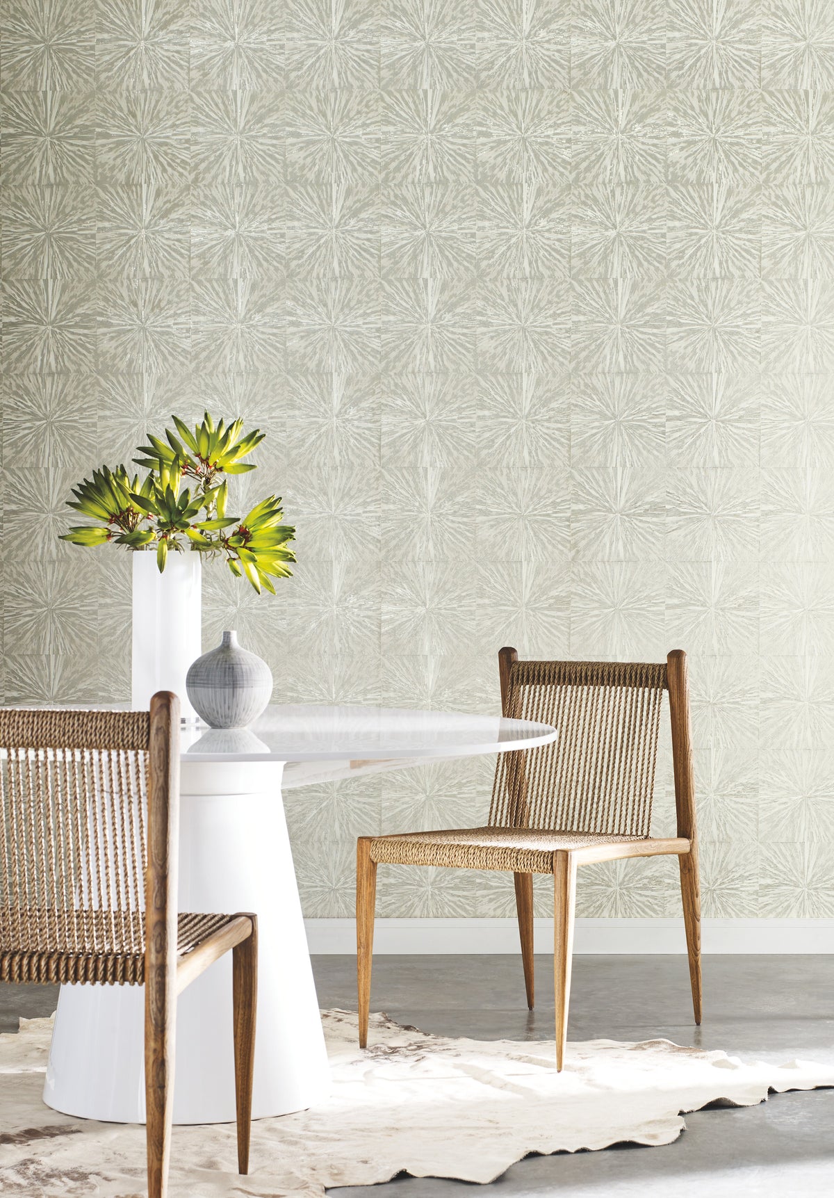 A modern dining area with two woven chairs, a round white table topped with a vase of green flowers, York Wallcoverings’ Squareburst Wallpaper on the wall, and a cowhide rug on the floor.