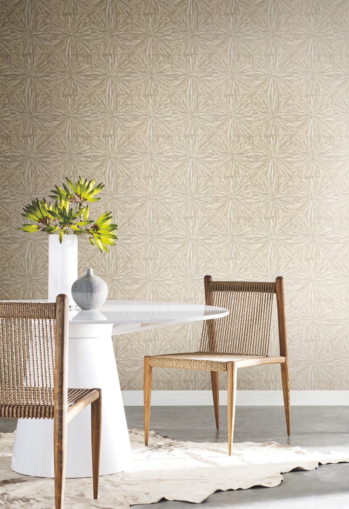 A modern dining space featuring a round white table, two woven wooden chairs, a gray vase, and a white vase with green leaves, arranged on a cowhide rug against a York Wallcoverings Squareburst Wallpaper accent wall.