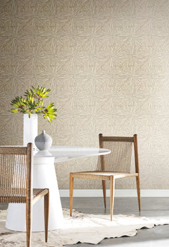 A modern dining space featuring a round white table, two woven wooden chairs, a gray vase, and a white vase with green leaves, arranged on a cowhide rug against a York Wallcoverings Squareburst Wallpaper accent wall.