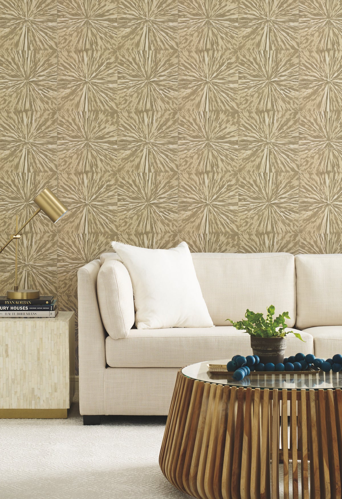 A modern living room featuring a beige sofa, white pillow, York Wallcoverings’ Squareburst Wallpaper, gold lamp, square side table, wooden coffee table, green plant, and decorative beads.