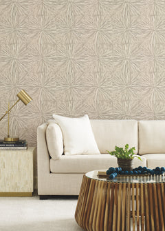 A beige sofa with white pillows sits beside a side table topped with a gold lamp and books. In front is a round wooden coffee table adorned with plants and decor. The walls feature York Wallcoverings’ Squareburst Wallpaper, showcasing its distinctive textured, starburst pattern.