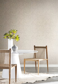 A modern dining area with a round white table, two wooden chairs featuring woven seats, a gray vase, a white vase with green foliage, and York Wallcoverings’ Squareburst Wallpaper adding a light pattern to the walls. A cowhide rug lies beneath the table.