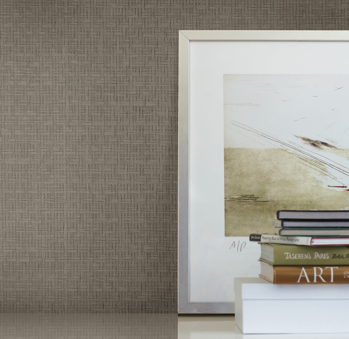 A framed abstract art piece sits on a white surface next to a small stack of books, set against a wall covered in Candice Olson Tatami Weave Wallpaper by York Wallcoverings.