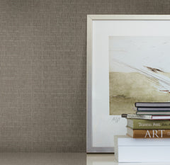 A framed abstract art piece sits on a white surface next to a small stack of books, set against a wall covered in Candice Olson Tatami Weave Wallpaper by York Wallcoverings.