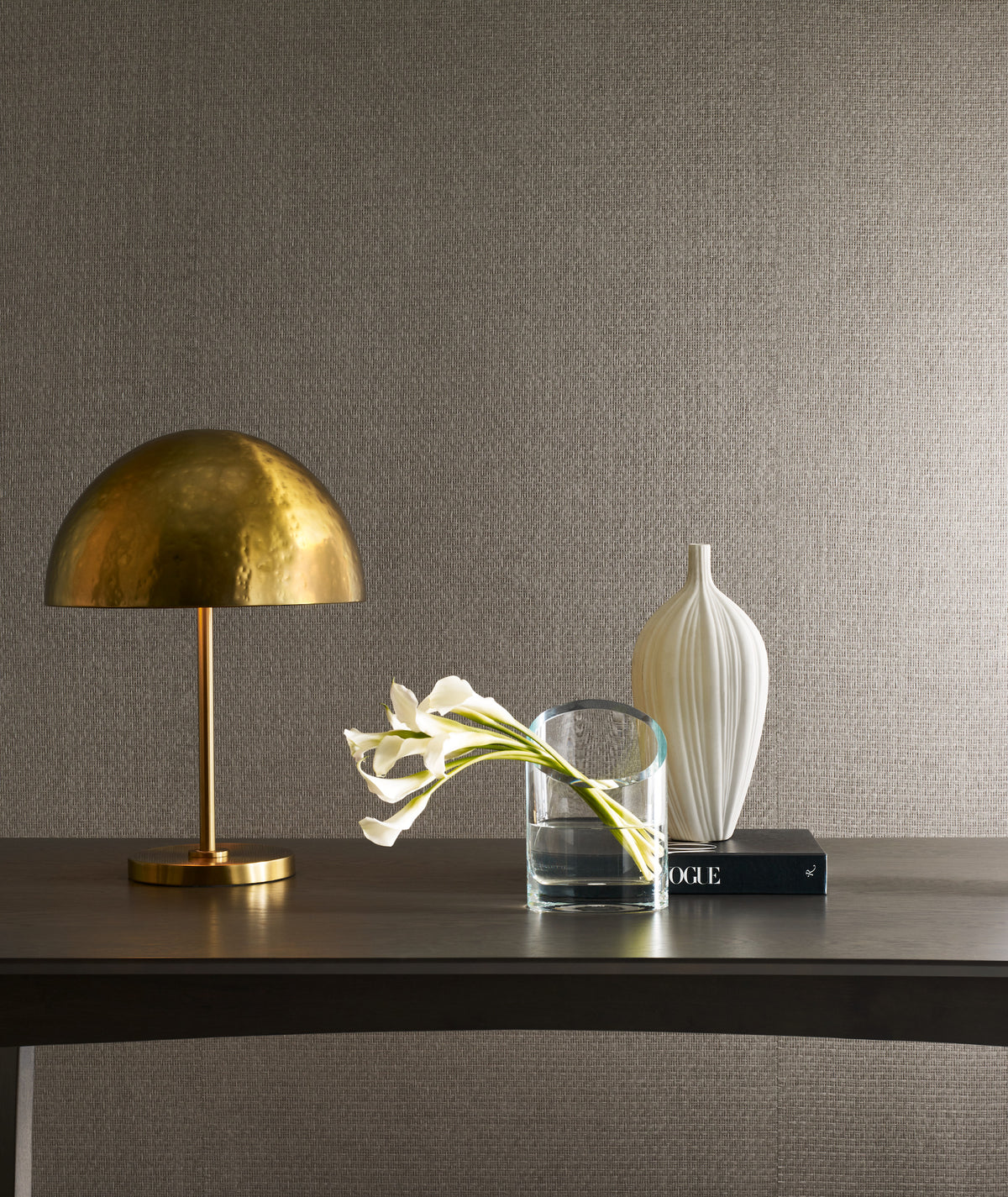A brass dome lamp, a clear glass vase with white calla lilies, and a white ceramic vase are arranged on a dark table against a wall adorned with Candice Olson Tatami Weave Wallpaper by York Wallcoverings. A black book rests beneath the white vase.