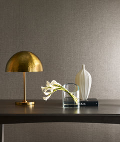 A brass dome lamp, a clear glass vase with white calla lilies, and a white ceramic vase are arranged on a dark table against a wall adorned with Candice Olson Tatami Weave Wallpaper by York Wallcoverings. A black book rests beneath the white vase.