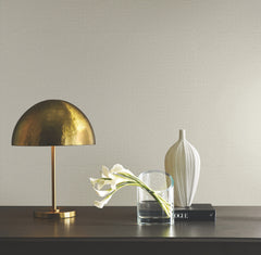 A brass dome lamp, a clear glass with calla lilies, a ribbed white vase, and a book sit on a dark wooden surface against a wall covered in Ronald Redding Tatami Weave Wallpaper by York Wallcoverings.
