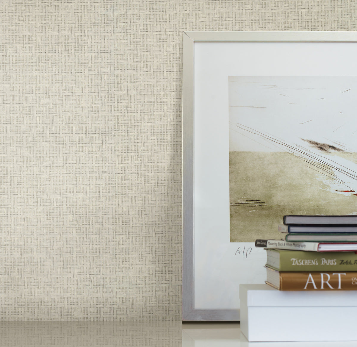 A framed abstract artwork leans against a wall covered in Candice Olson Tatami Weave Wallpaper by York Wallcoverings, near a stack of books and a white box on a glossy surface. Only part of the art and book titles are visible.