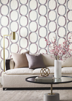 A cream-colored sofa sits in front of a wall covered in Candice Olson Interlock Wallpaper, which features a repeating pattern of interlocking purple and gray ovals.